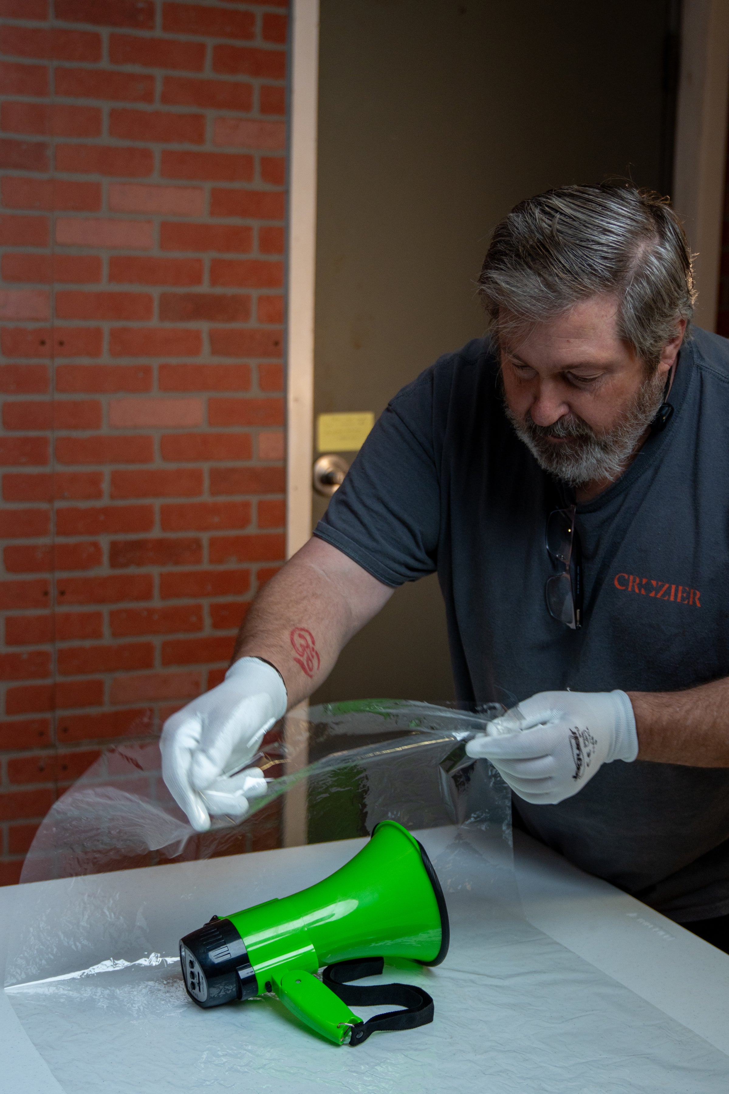 Man in gray shirt with gloves handling a green megaphone on a table against a brick wall.